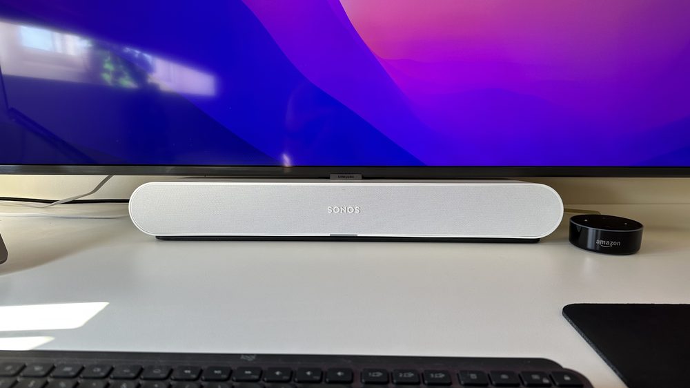 A Sonos Ray soundbar on a desk beneath a monitor, fed from a Mac
                by an optical cable.